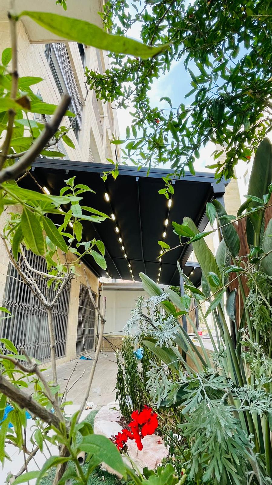 פרגולת אלומיניום מוקפת צמחייה ועצי רימון פורחים